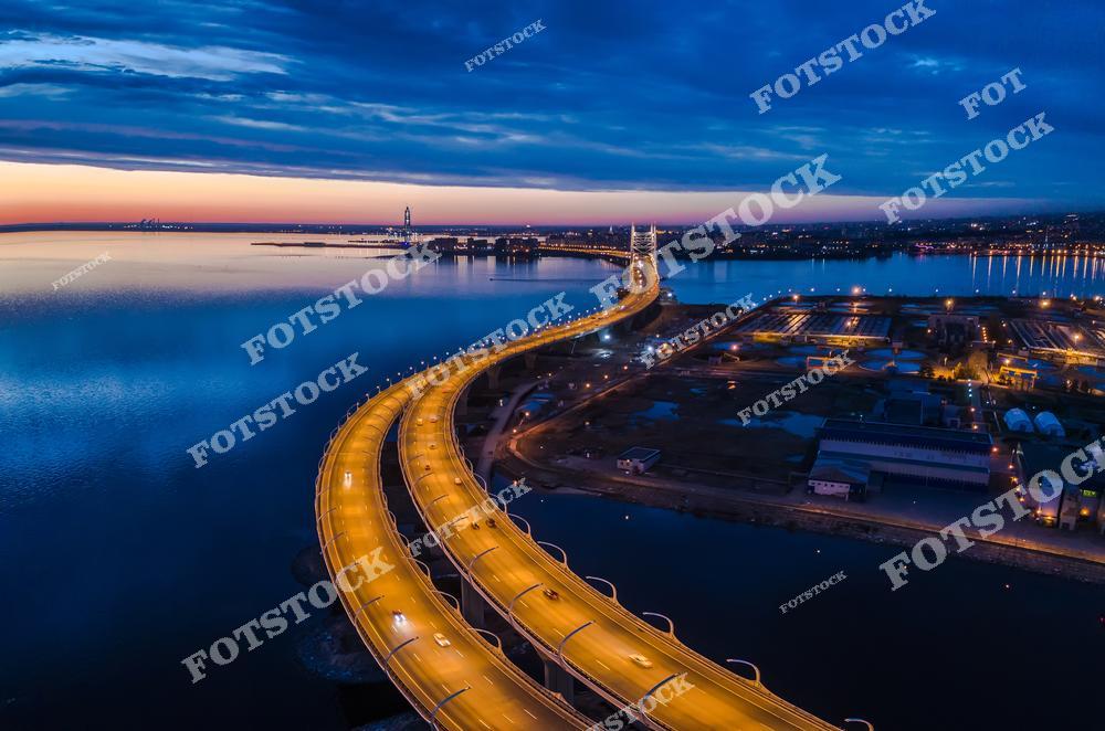 Канонерский остров. Панорамный вид на ночной Санкт-Петербург с высоты, где светящиеся огни набережной отражаются в воде Невы во время белых ночей. Архитектура зданий, подсветка мостов и атмосферная дымка создают романтичный и выразительный городской пейзаж северной столицы России. Фотография передаёт уникальное настроение белых ночей, красоту городской инфраструктуры и величие исторического центра Петербурга. Изображение идеально подходит для публикаций о путешествиях по России, ночных панорамах города, архитектуре Санкт-Петербурга и туристических достопримечательностях.